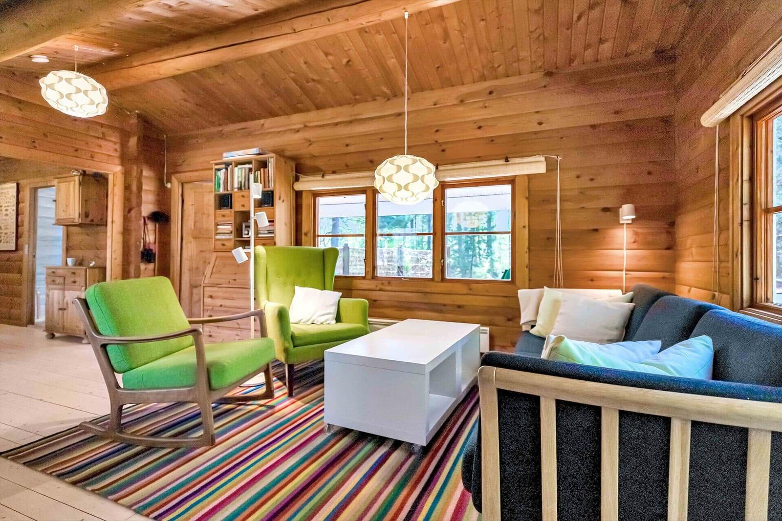 Wohnzimmer mit Holzwand, grünen Sesseln, Sofa und buntem Teppich.