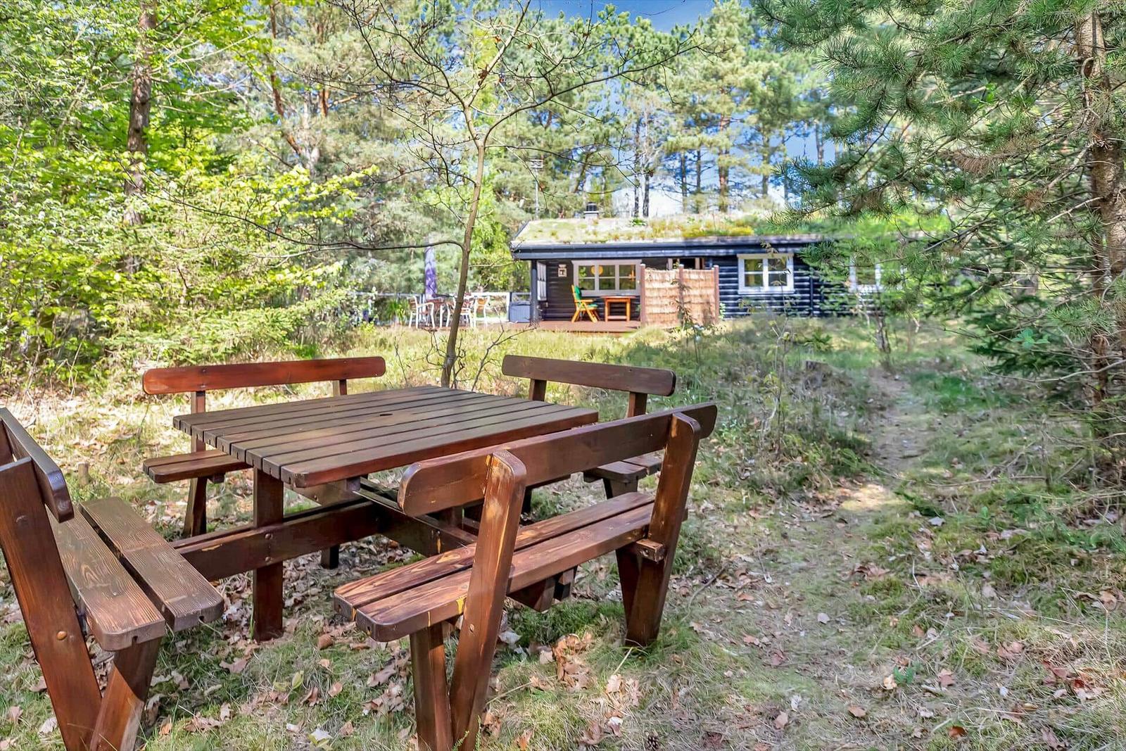 Holztisch mit Bänken im Wald. Im Hintergrund ein Holzhaus mit Dachboden.