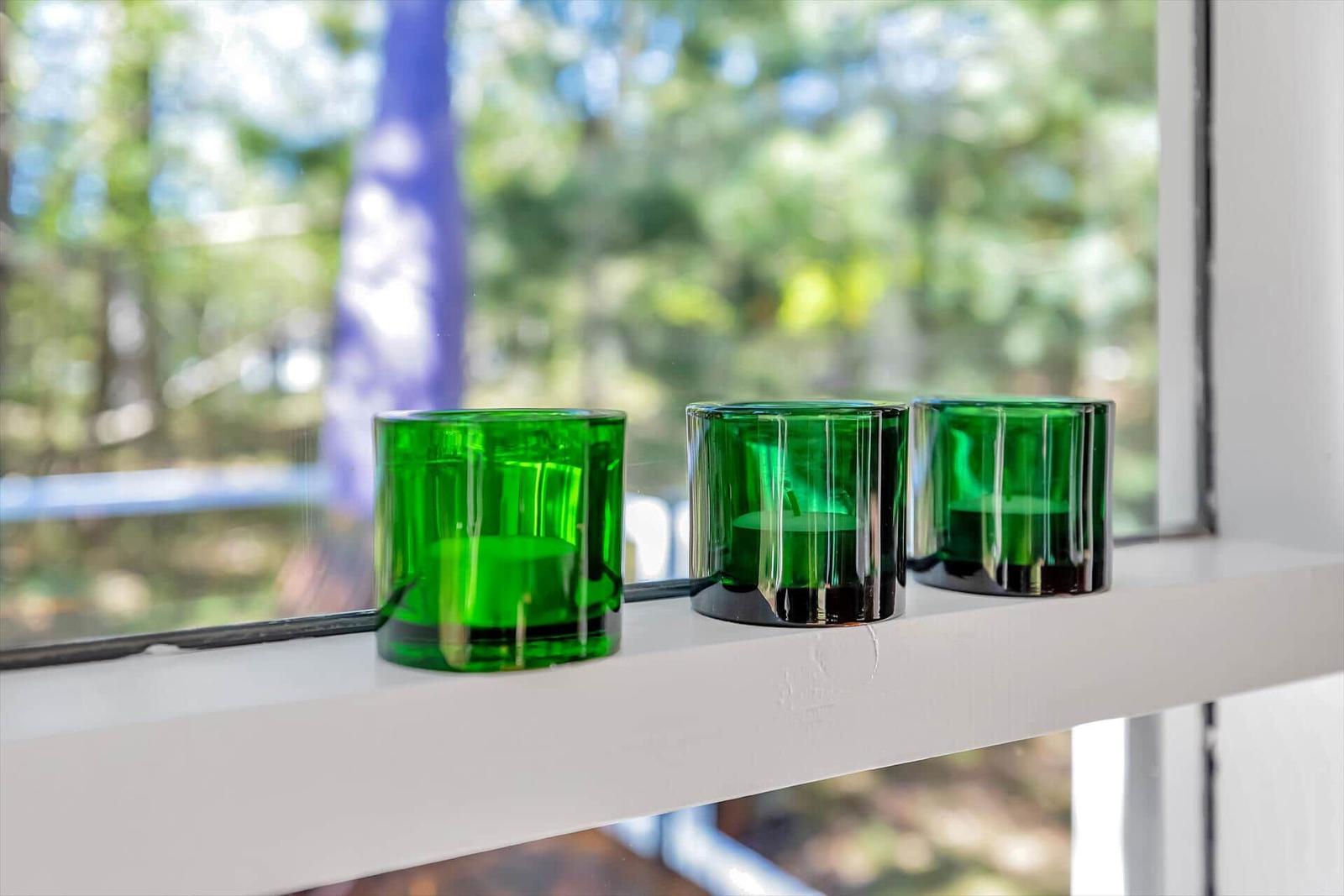 Drei grüne Teelichter stehen auf einem weißen Fensterbrett vor einer verschwommenen grünen Landschaft.