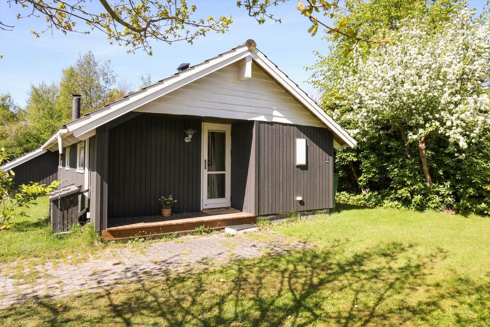 Holzhaus mit weißem Dach und Eingangstür. Grün umgeben mit blühendem Baum.