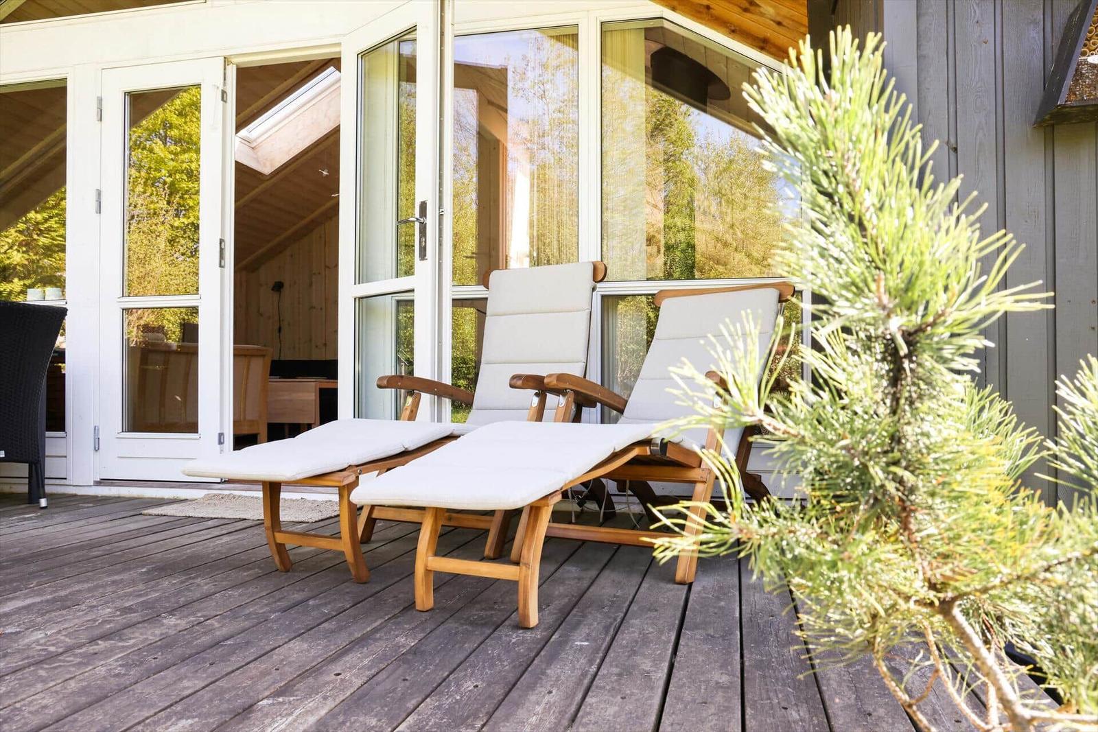 Zwei Liegestühle auf Holzterrasse vor Glasfront mit Blick ins Haus.