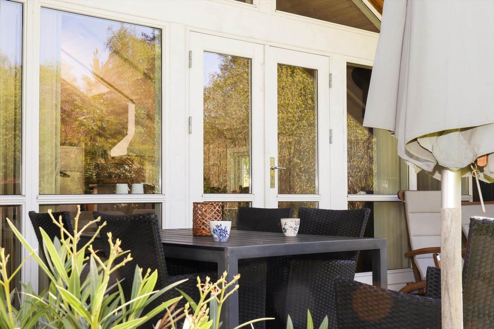 Terrasse mit Tisch, Stühlen und Sonnenschirm. Hintergrund: Glasfront und Pflanzen.