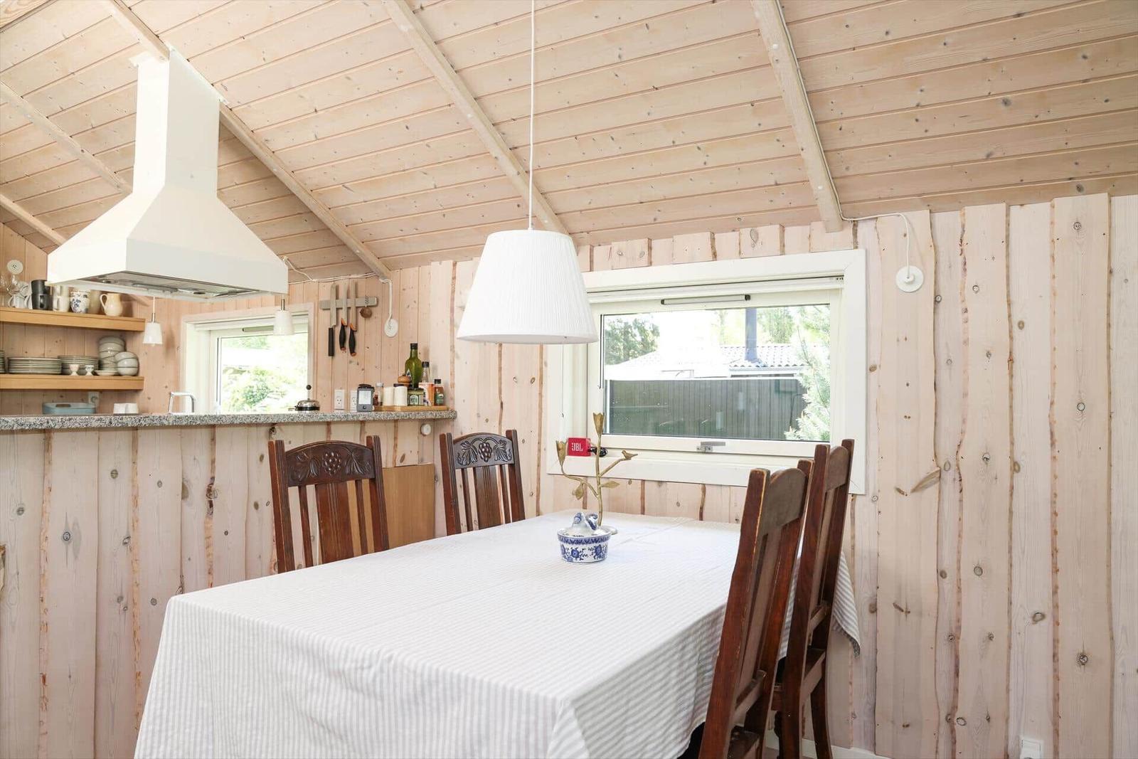 Esszimmer mit Holzwand, Tisch und Stühlen. Fenster und Küchenbereich sichtbar.