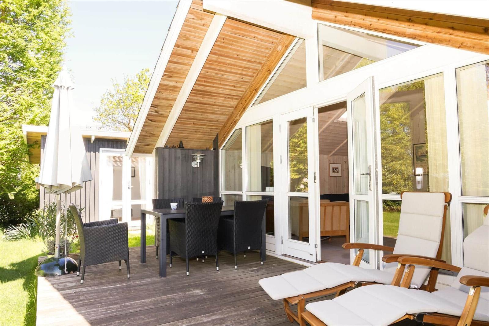 Terrasse mit Tisch, Stühlen und Liegestühlen unter Holzdecke. Glastüren führen ins Haus.
