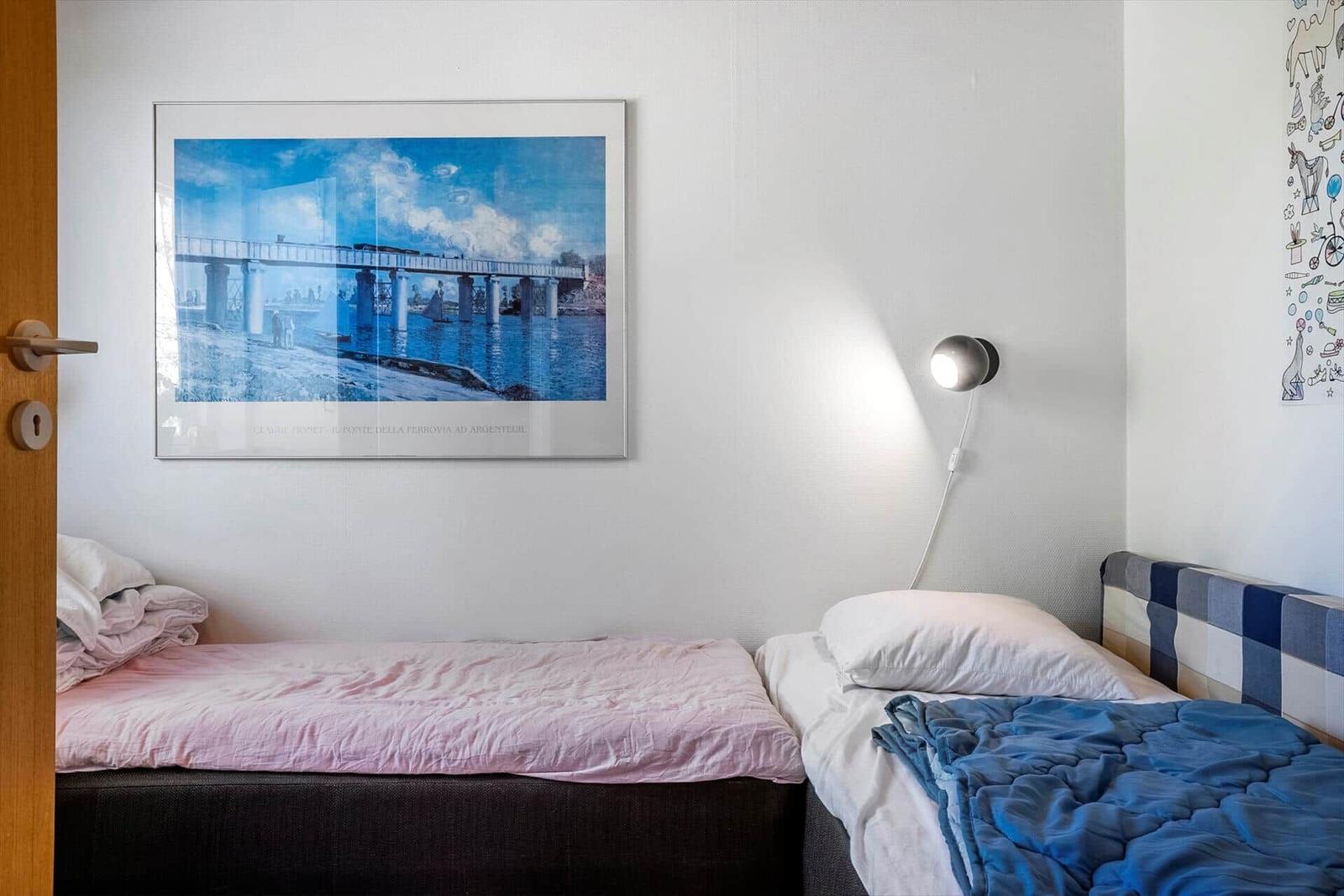 Two beds with pink and blue bedding, wall art, and a wall lamp.