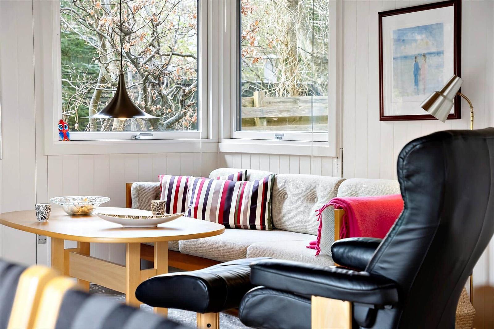 Ein Wohnzimmer mit Sofa, Tisch und schwarzer Ledercouch. Durch Fenster blickt man auf Bäume.