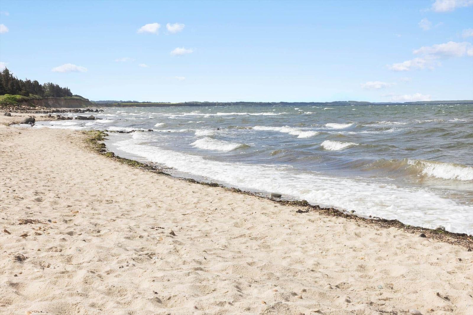 Strand mit Sand und Wellen, Küstenlinie mit Felsen und Bäumen im Hintergrund.