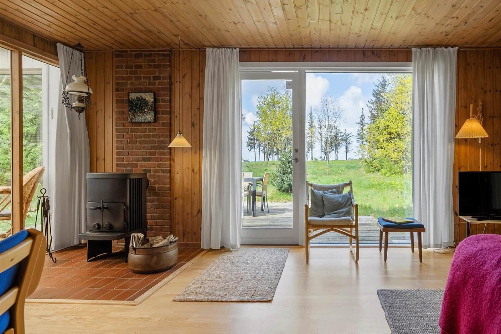 Wohnraum mit Holzwand, Kaminofen und großem Fensterblick auf Grünland.