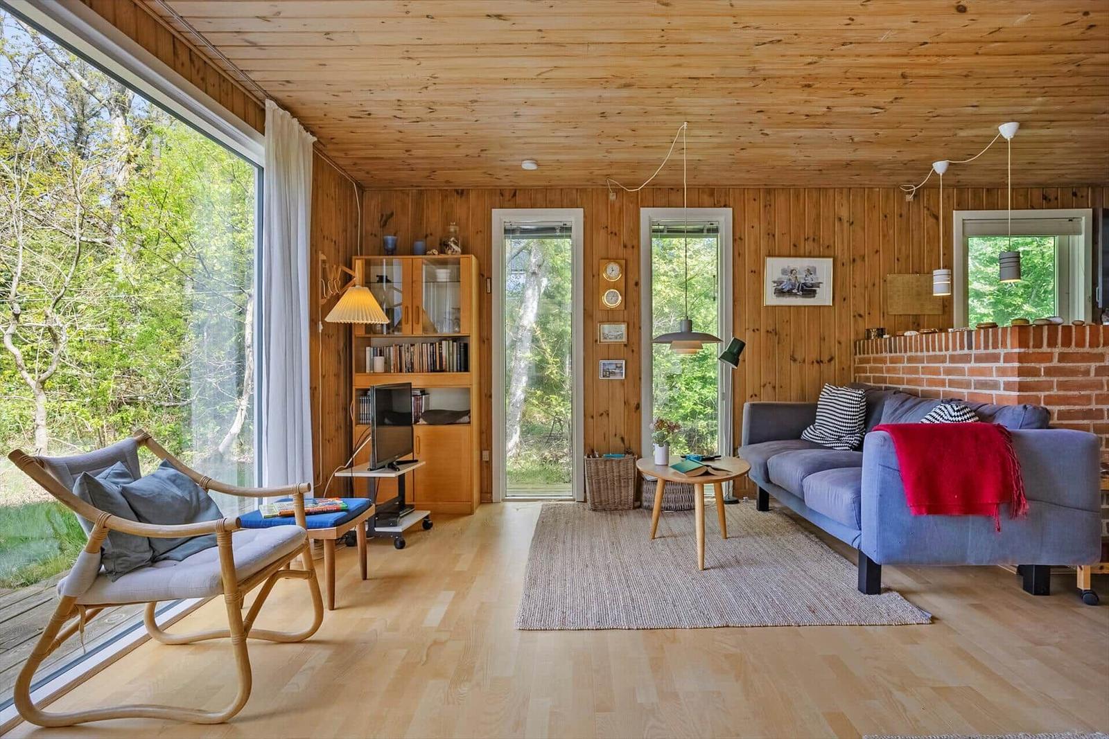Wohnzimmer mit Holzdecke, Sofa, Sessel und großem Fenster zur Natur.