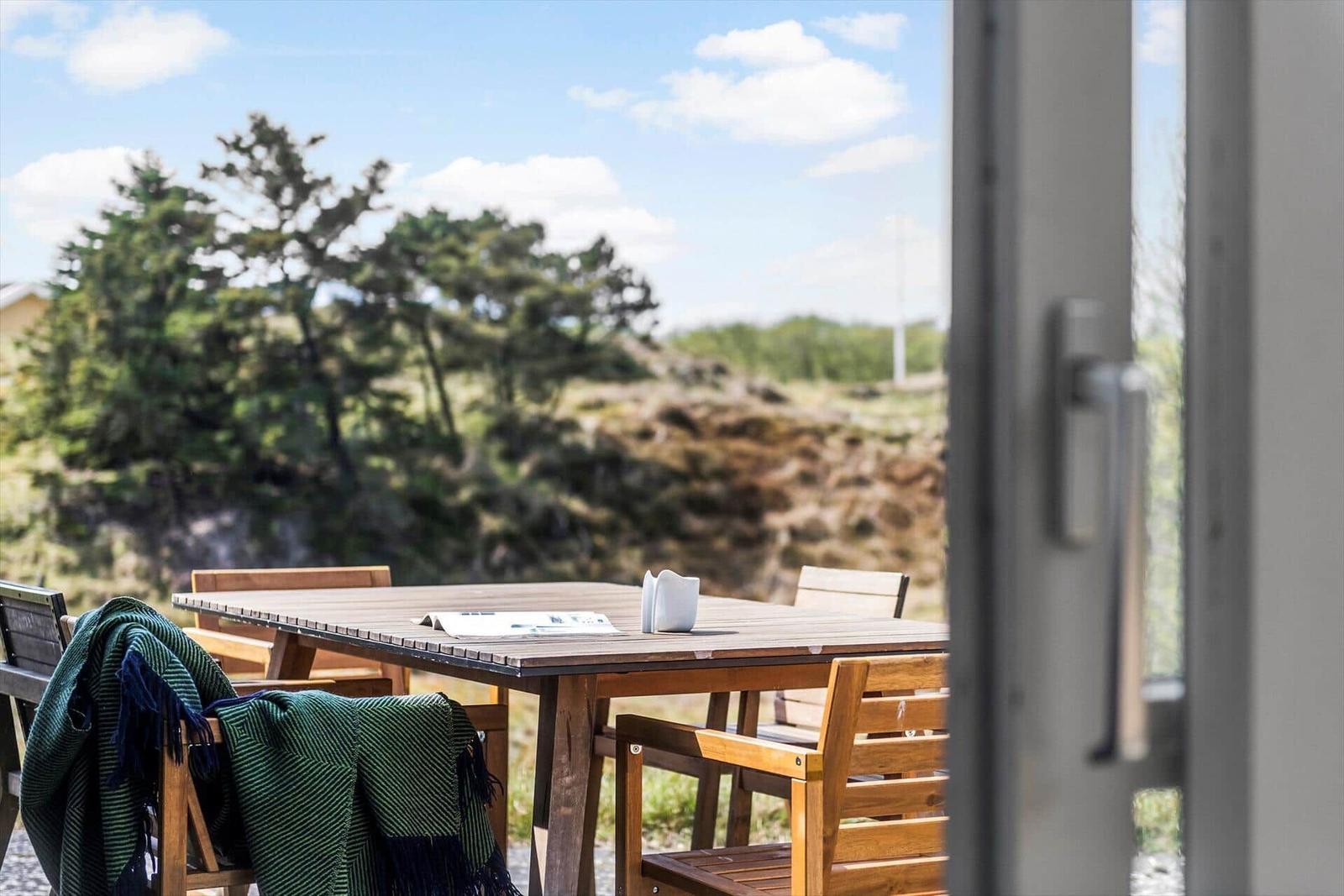Terrasse mit Holztisch und Stühlen. Grüner Schal liegt auf einem Stuhl. Hintergrund: Bäume und Himmel.