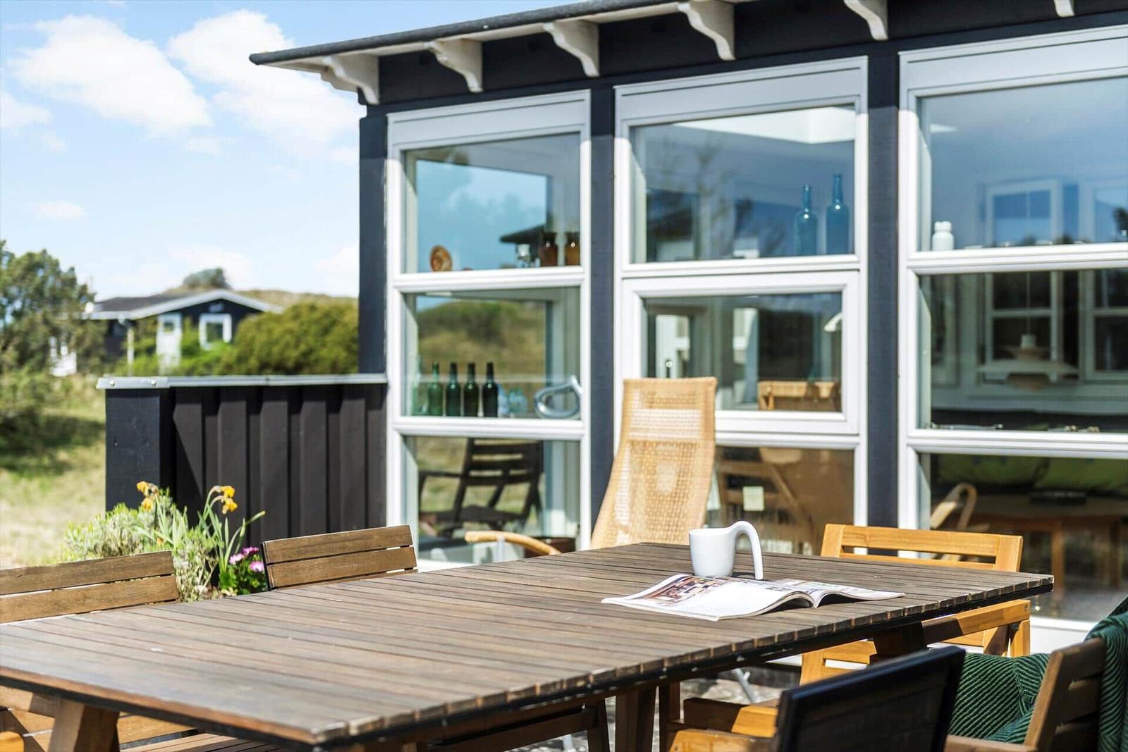 Terrasse mit Holztisch und Stühlen. Im Hintergrund ein Haus mit großen Fenstern.
