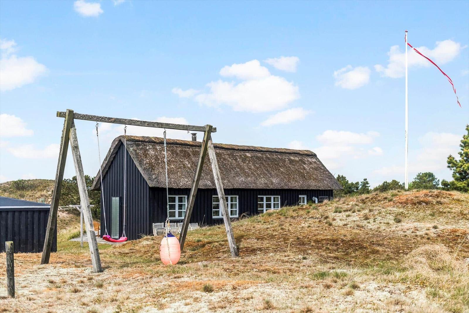 Schwarzes Haus mit strohgedecktem Dach, Spielgeräte und Flagge auf grüner Wiese.