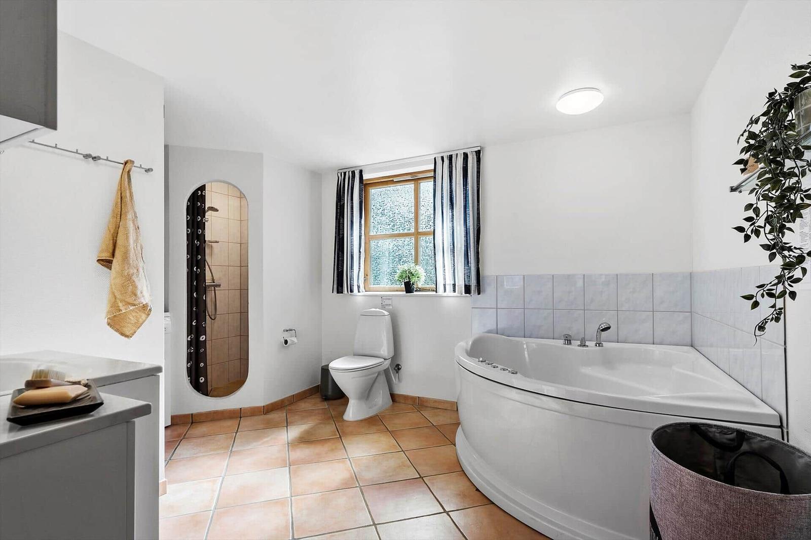 Bad mit Badewanne, Toilette und Dusche. Fenster mit Vorhängen und Pflanze.