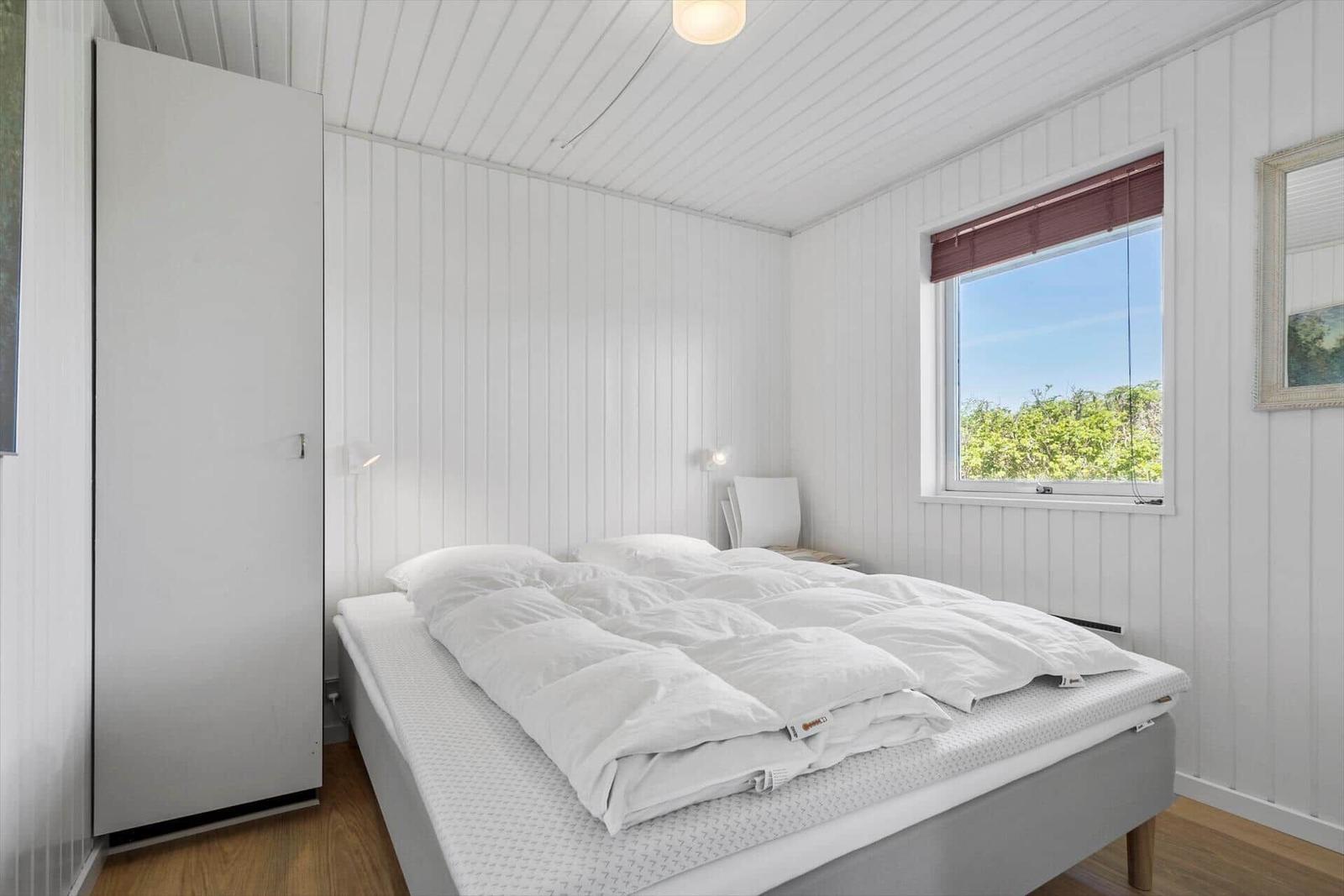 Schlafzimmer mit weißem Bett, Kleiderschrank und Fenster mit Blick auf Grün.