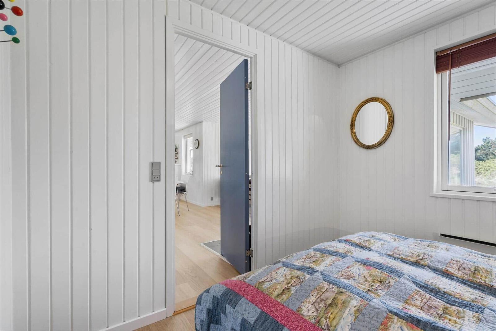 Schlafzimmer mit Bett, Fenster und Tür zum Flur. Wände sind weiß verkleidet.