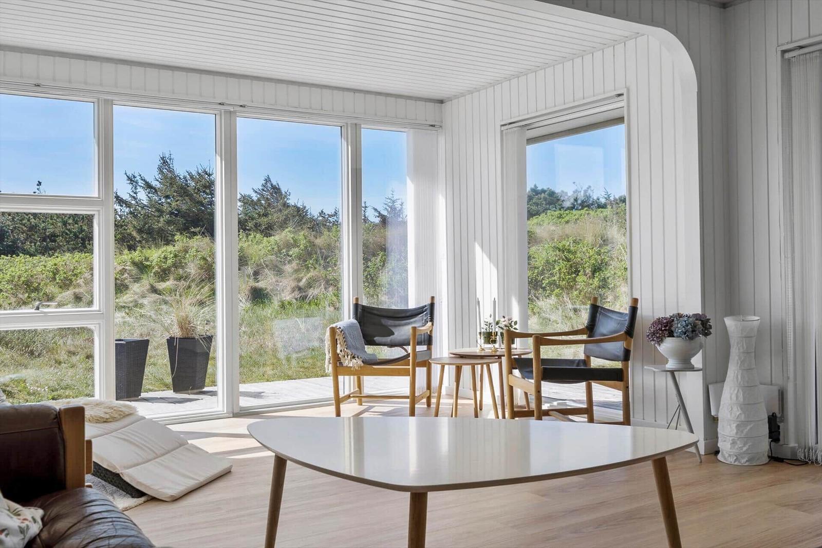 Lichter Raum mit großem Fensterblick auf Grünflächen und Bäume. Holztisch, zwei Stühle und Sofa im Vordergrund.