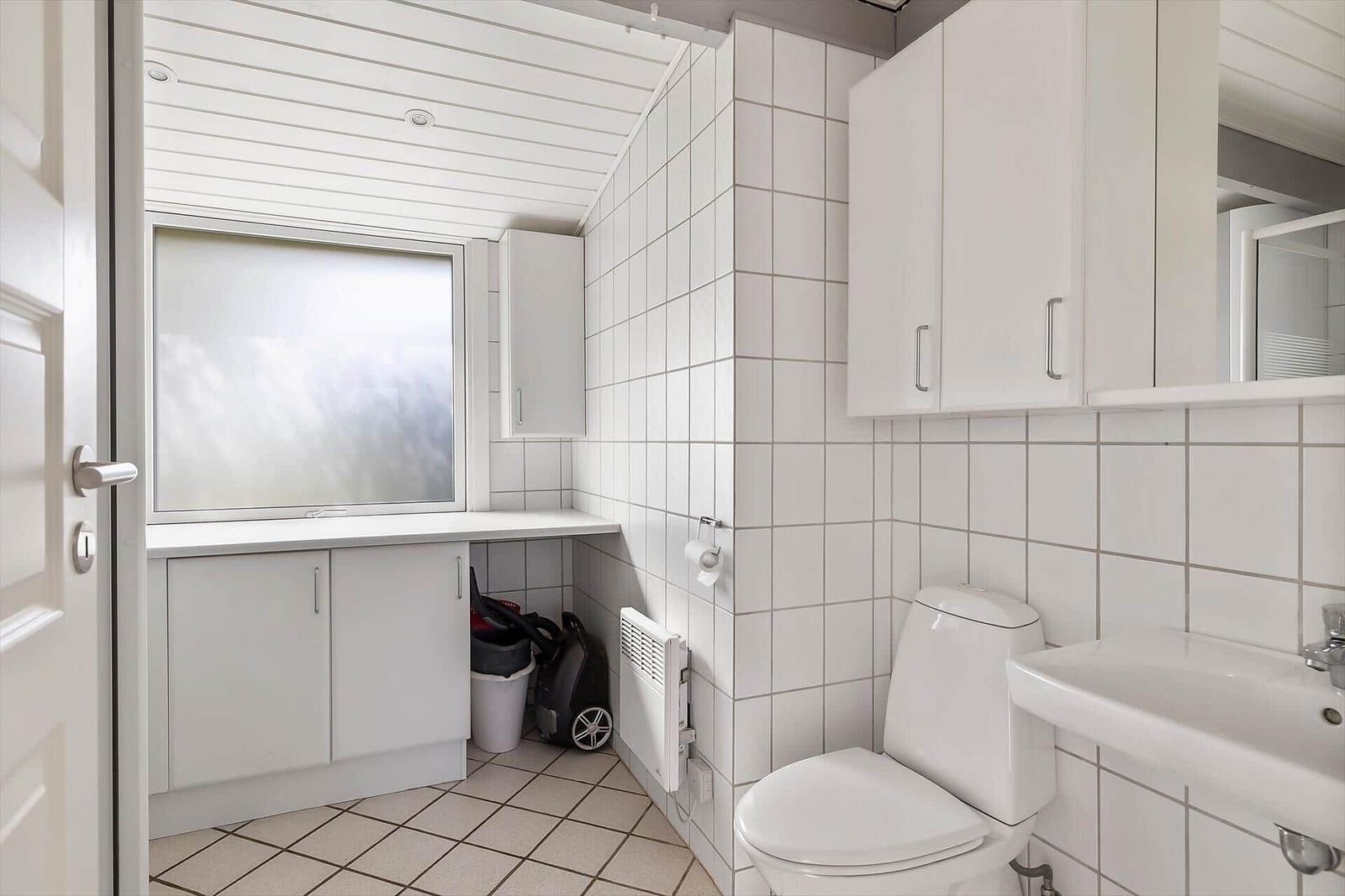 Badezimmer mit Toilette, Waschbecken und Fenster. Weiße Fliesen und Schränke.