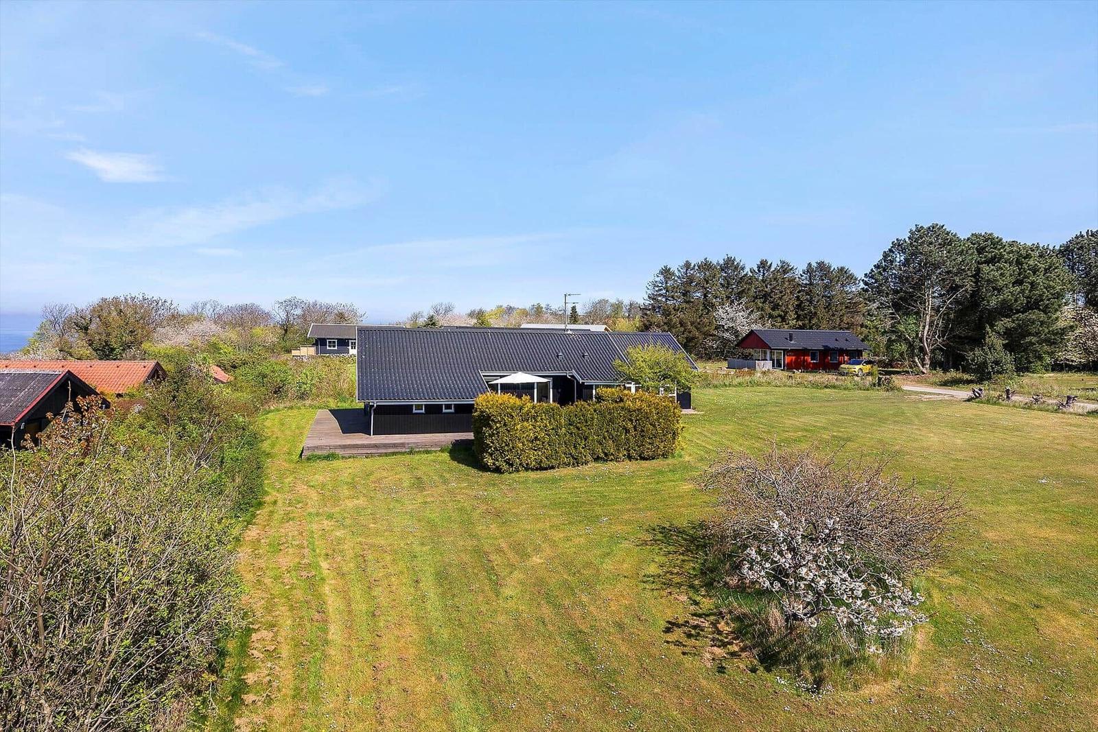 Modernes Haus mit Terrasse, umgeben von Grünfläche und Bäumen unter blauem Himmel.