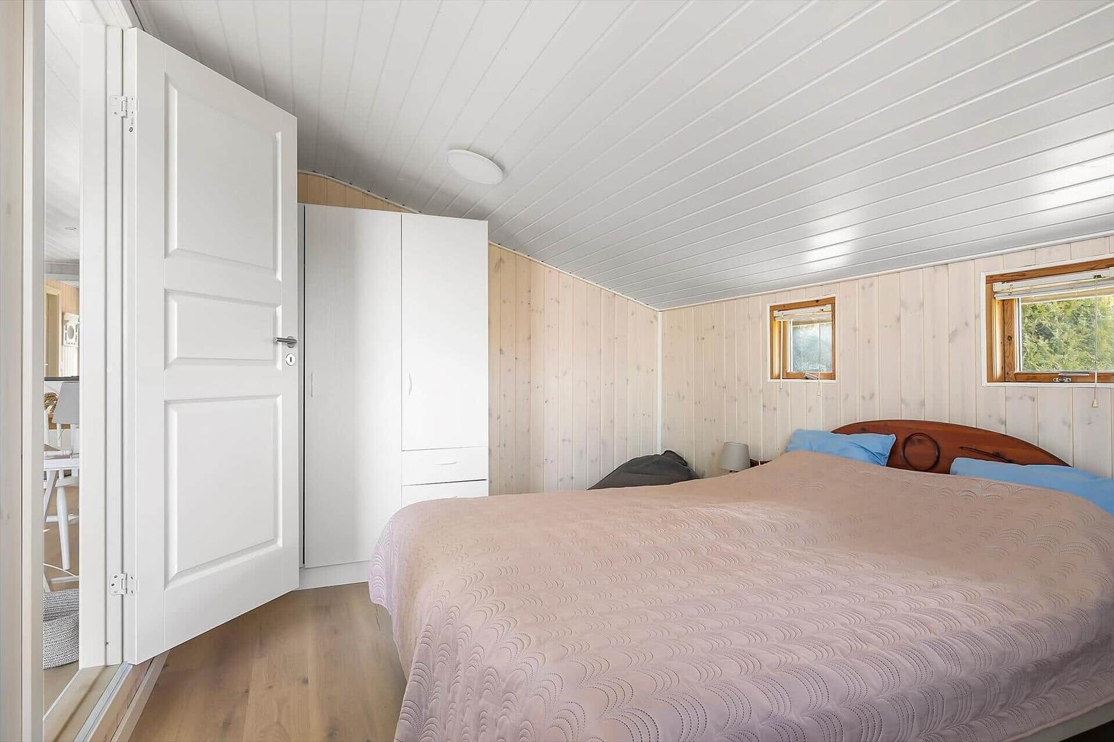 Schlafzimmer mit Bett, Kleiderschrank und Fenster. Holzwände und weißer Deckenverkleidung.