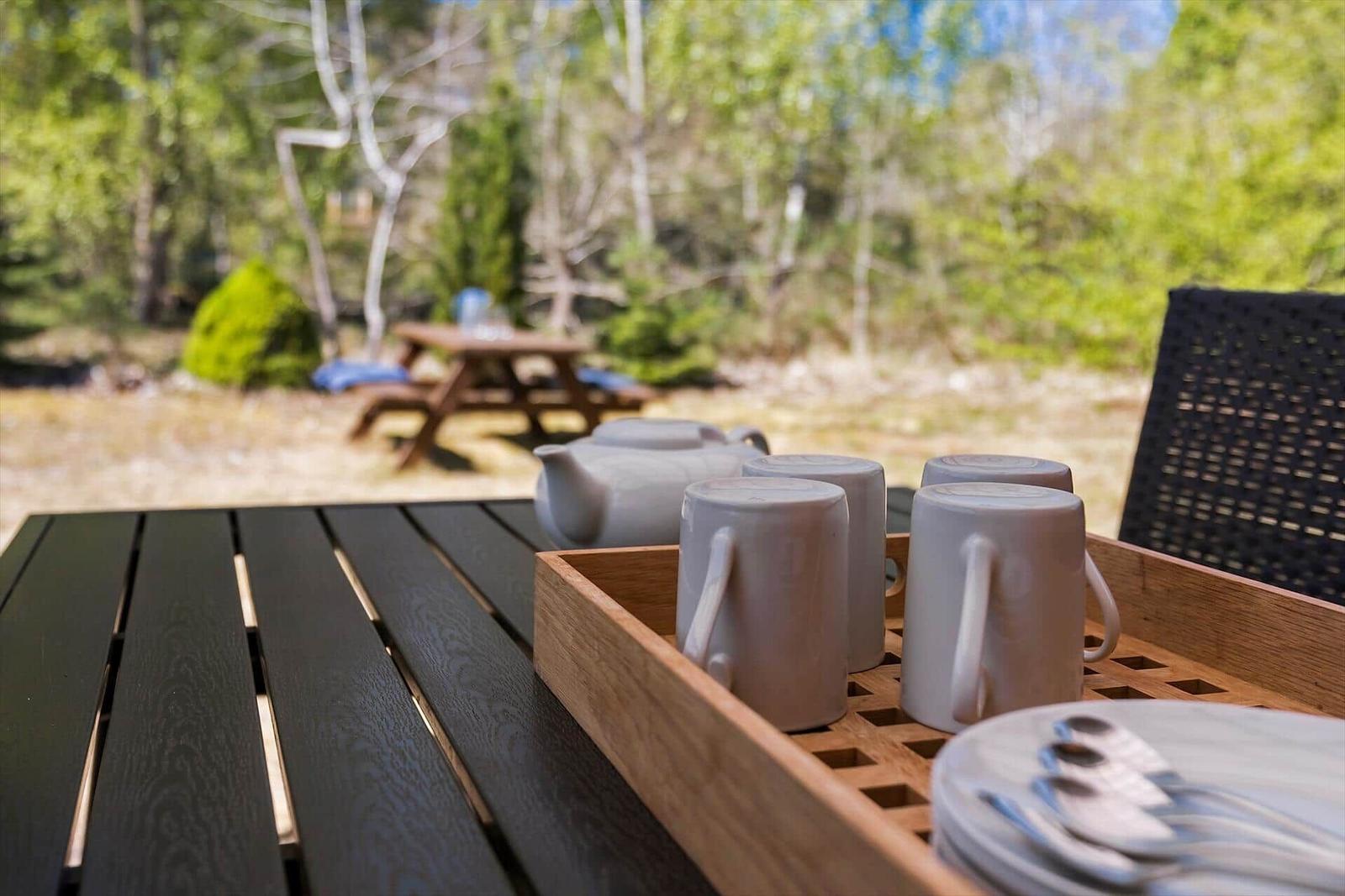 Außentisch mit Tee- und Geschirrset vor Wald im Hintergrund.