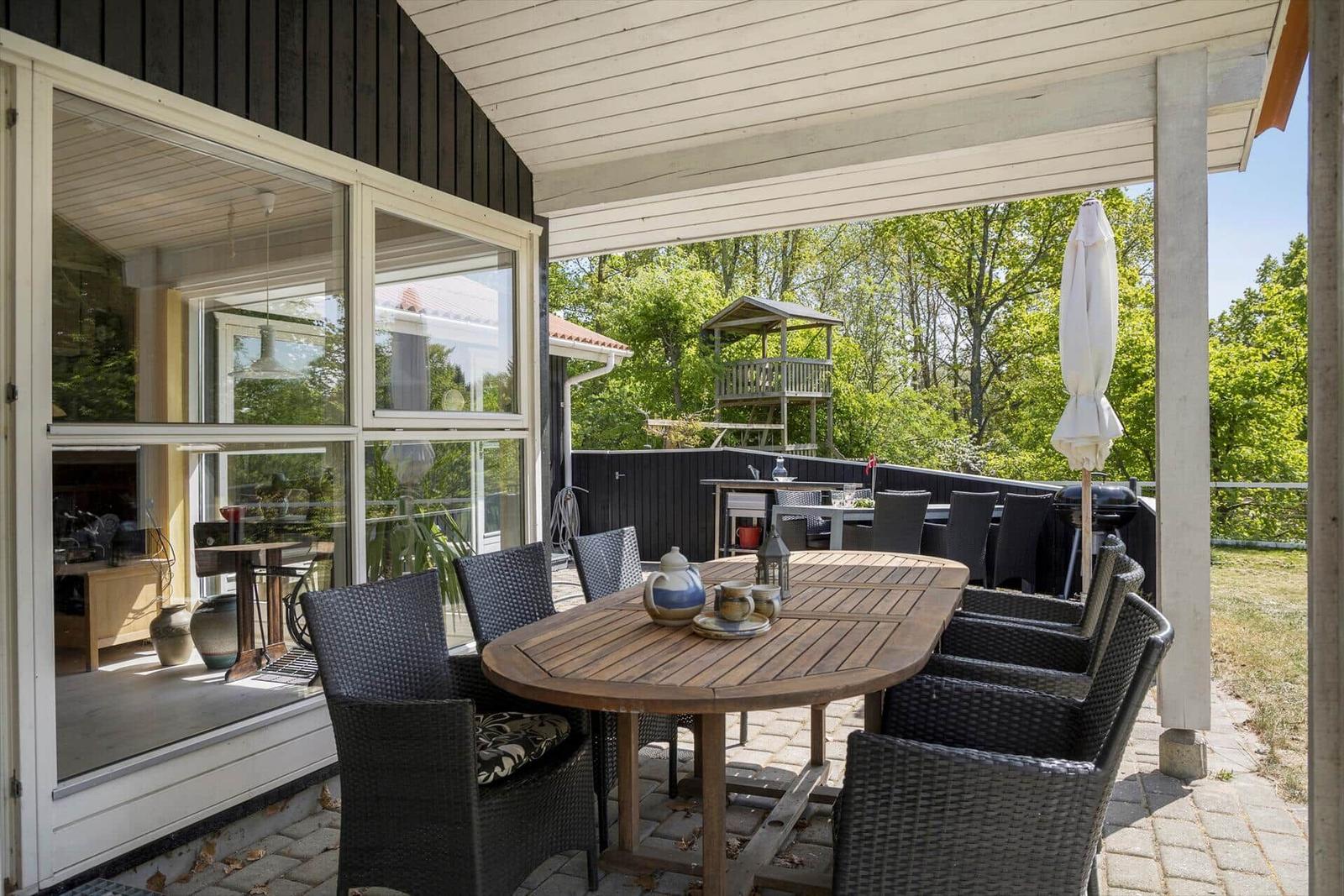 Terrasse mit Tisch, Stühlen und Sonnenschirm. Blick auf grüne Bäume und einen Spielplatz.