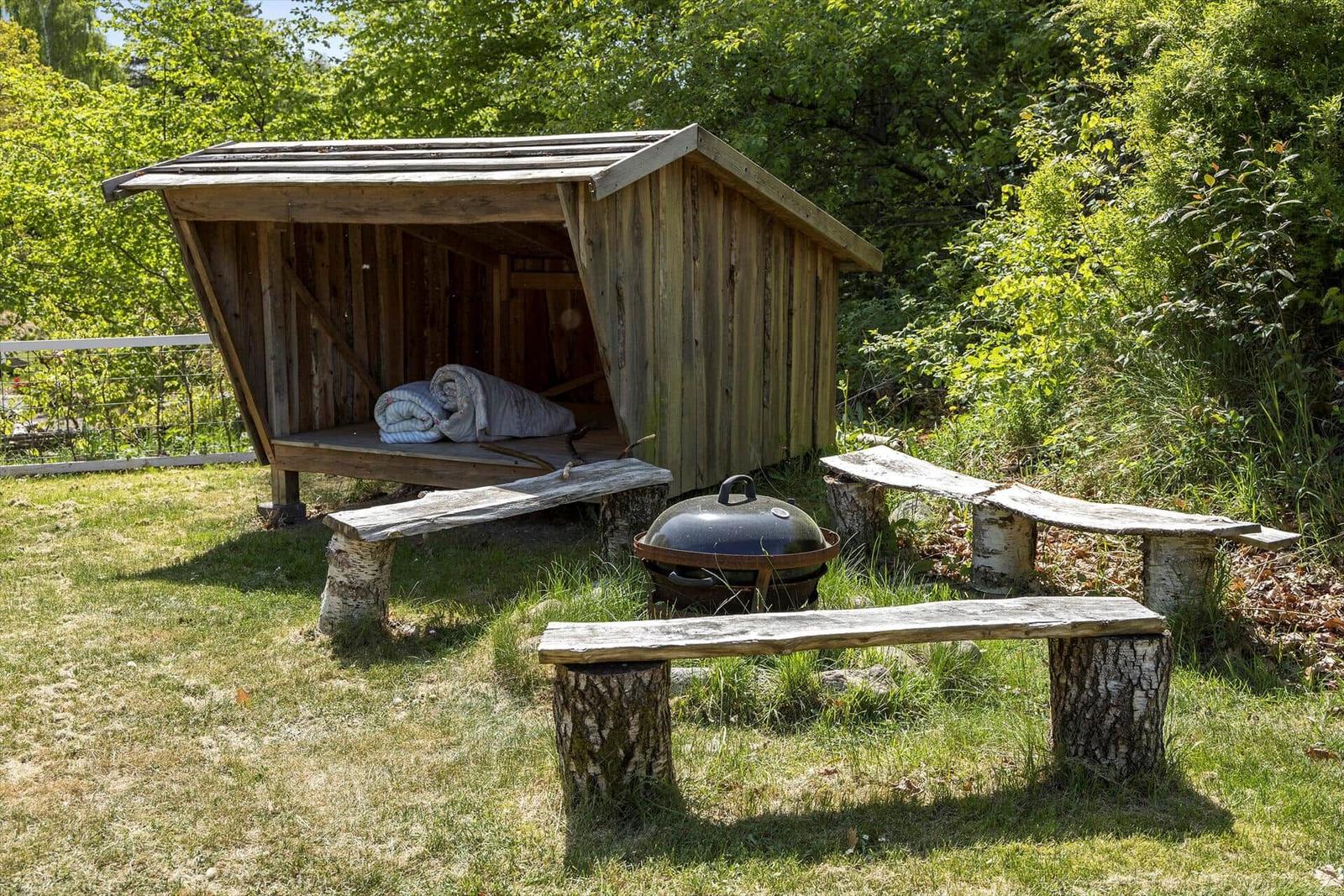 Holzschuppen mit Matratze, Grill und Bänken aus Baumstämmen im Garten.