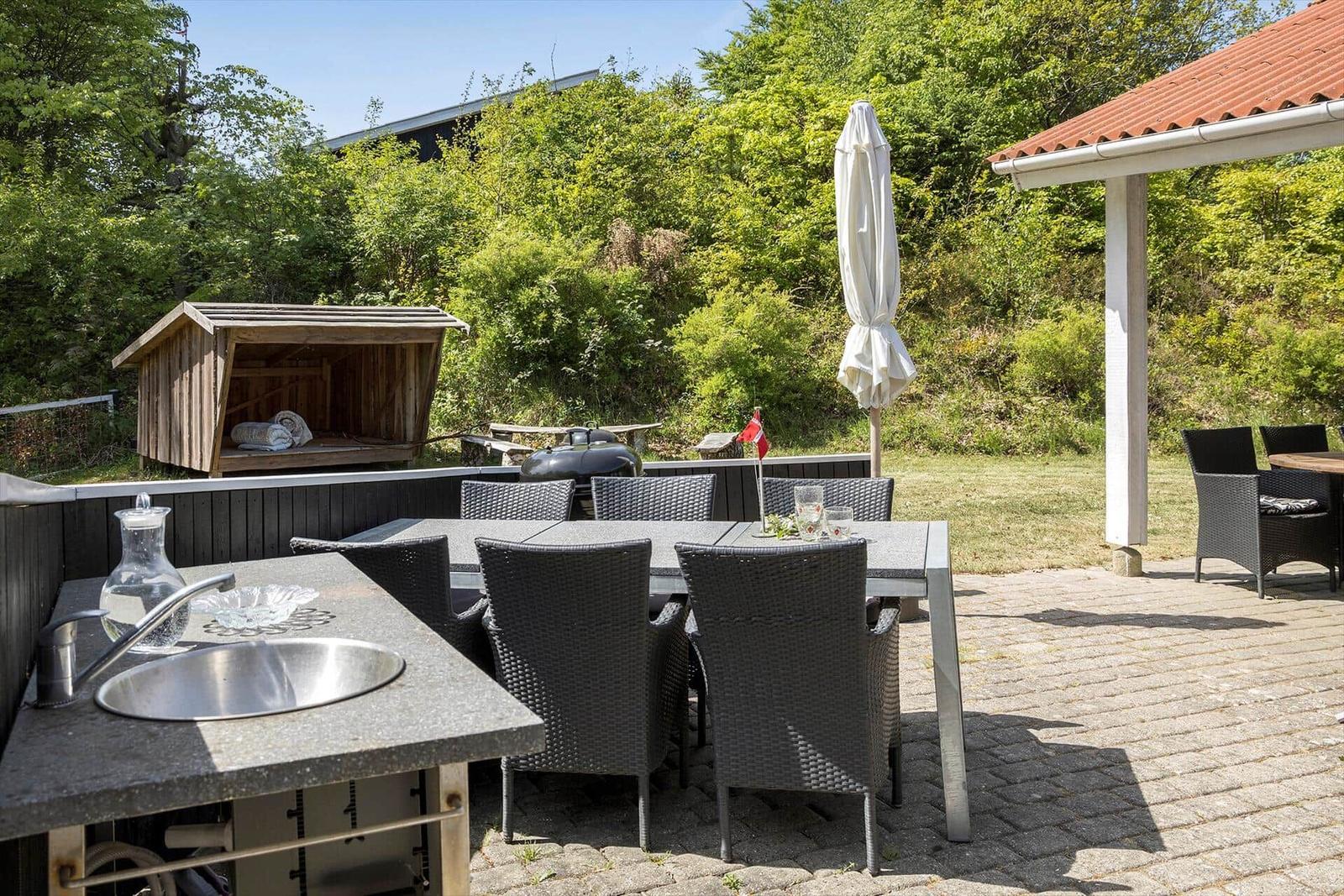 Außenbereich mit Grill, Sitzgruppe und Waschplatz. Hintergrund: Holzschuppen und grüne Vegetation.