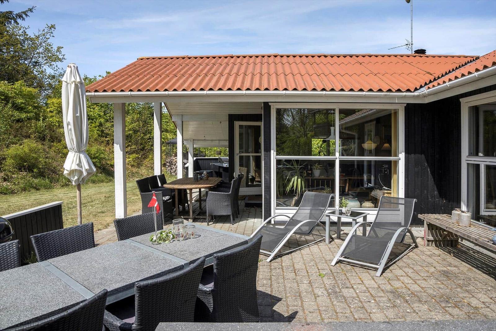 Terrasse mit Tischen, Stühlen und Liegen. Haus mit rotem Dach und Glasfront.
