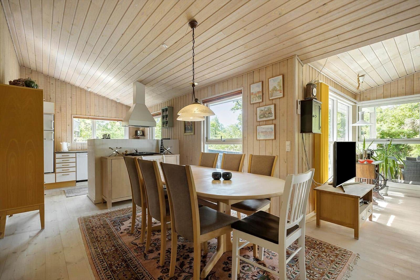 Ein Esszimmer mit Tisch, Stühlen und Küchenbereich. Holzwände und -decke. Fenster mit Blick auf Grün.