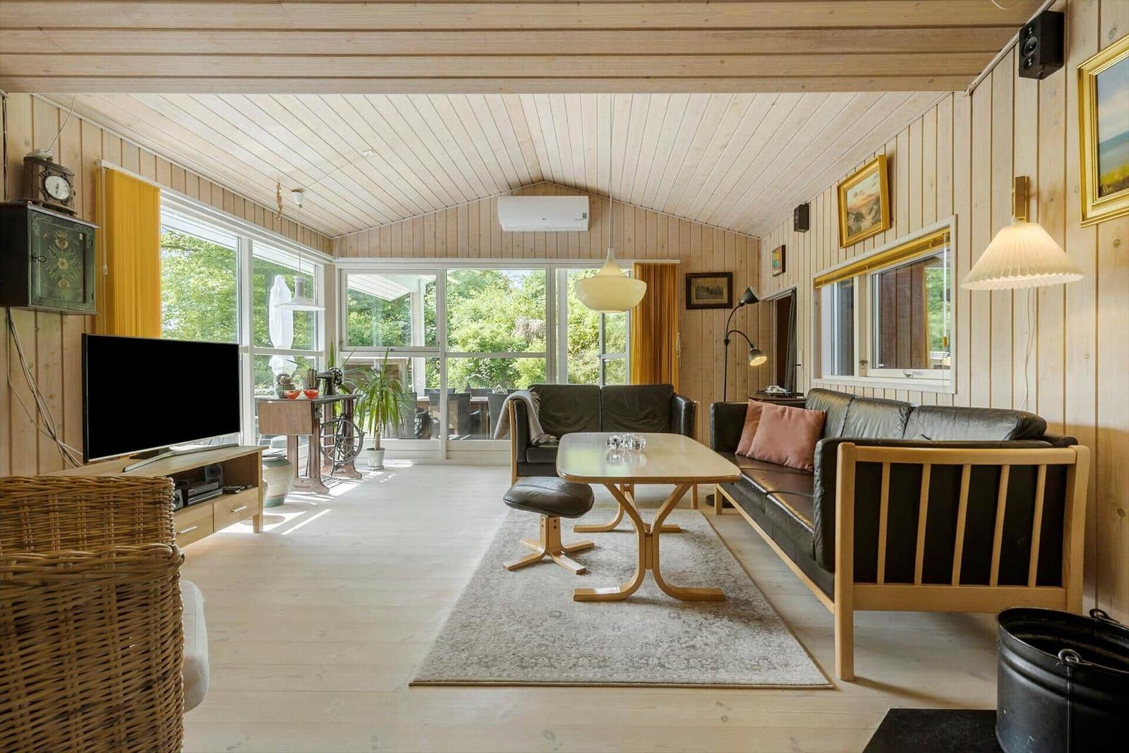 Wohnzimmer mit Holzwand, Sofa, Tisch und Fernseher. Große Fenster mit Blick auf Grün.