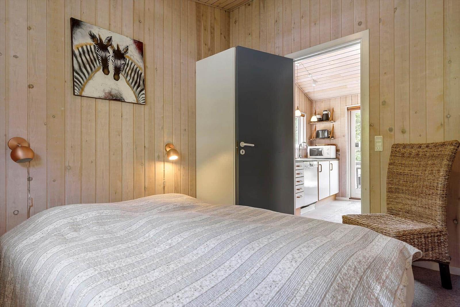 Schlafzimmer mit Bett, Wandbild und Stuhl. Küche im Hintergrund sichtbar.