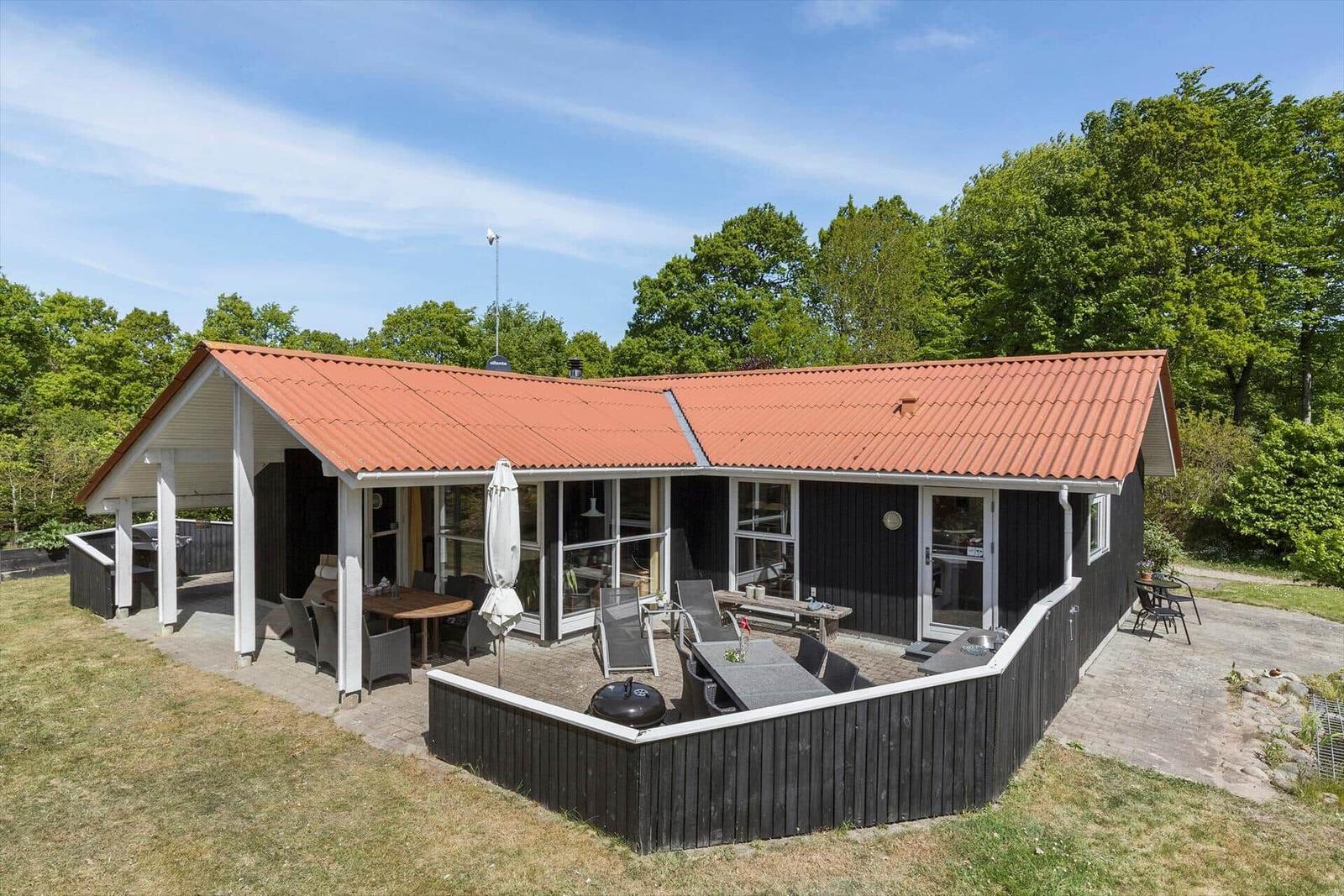 Haus mit Terrasse, Grill und Garten. Umgeben von Bäumen unter blauem Himmel.