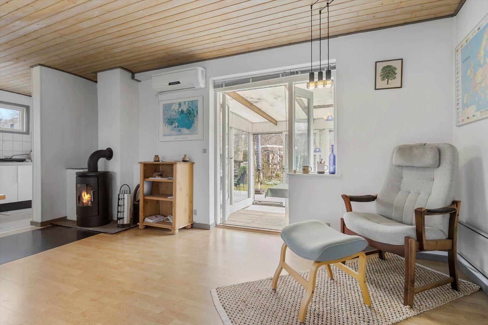 Living room with wooden ceiling, wood stove, and armchair. Door leads to terrace.