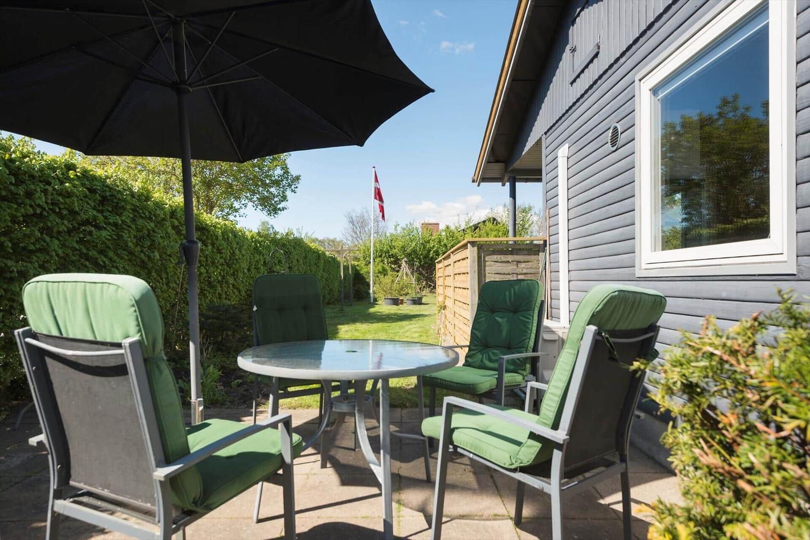 Terrasse mit Tisch, vier Stühlen und Sonnenschirm. Hintergrund: Grünhecke und dänische Flagge.