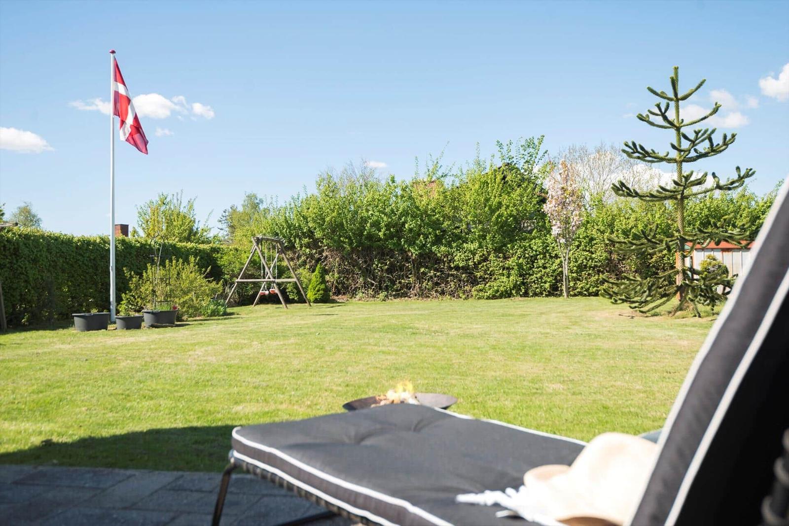 Garten mit Liegestuhl, Feuerkorb und dänischer Flagge. Hintergrund: Grasfläche, Spielgeräte und Bäume.