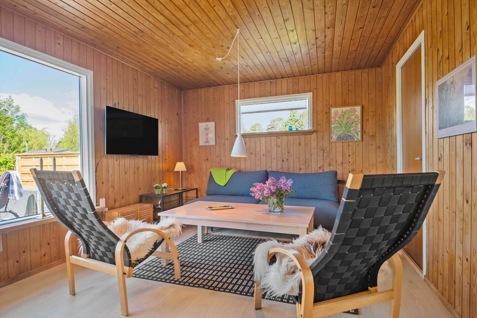 Wohnzimmer mit Holzwand, Sofa, zwei Stühlen und Fernseher. Fenster mit Blick auf Garten.