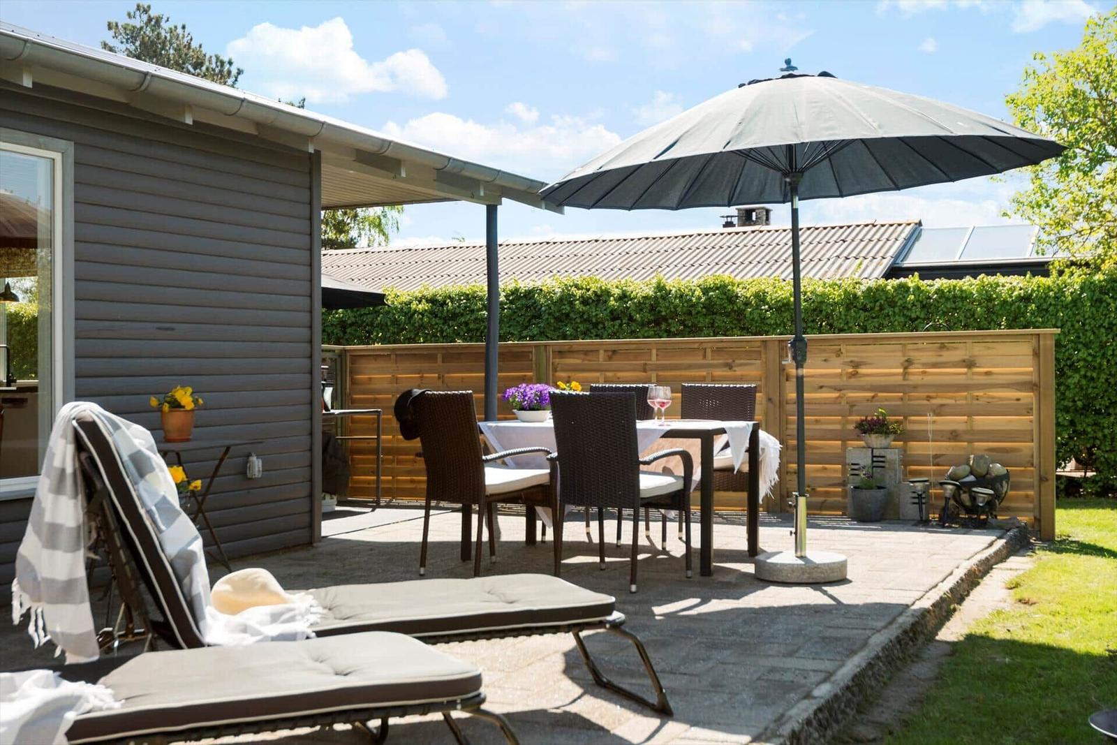 Terrasse mit Liegestühlen, Tisch und Sonnenschirm. Hinterhof mit Holzzaun und Grünflächen.