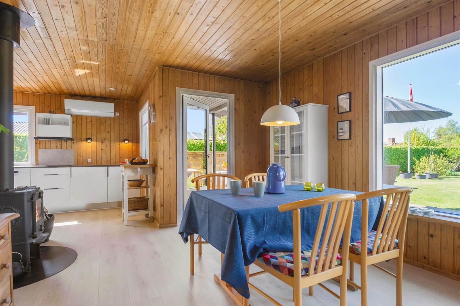 Küche und Essbereich mit Holzwand und -decke. Tisch mit blauer Tischdecke und Stühlen. Fenster mit Gartenblick.