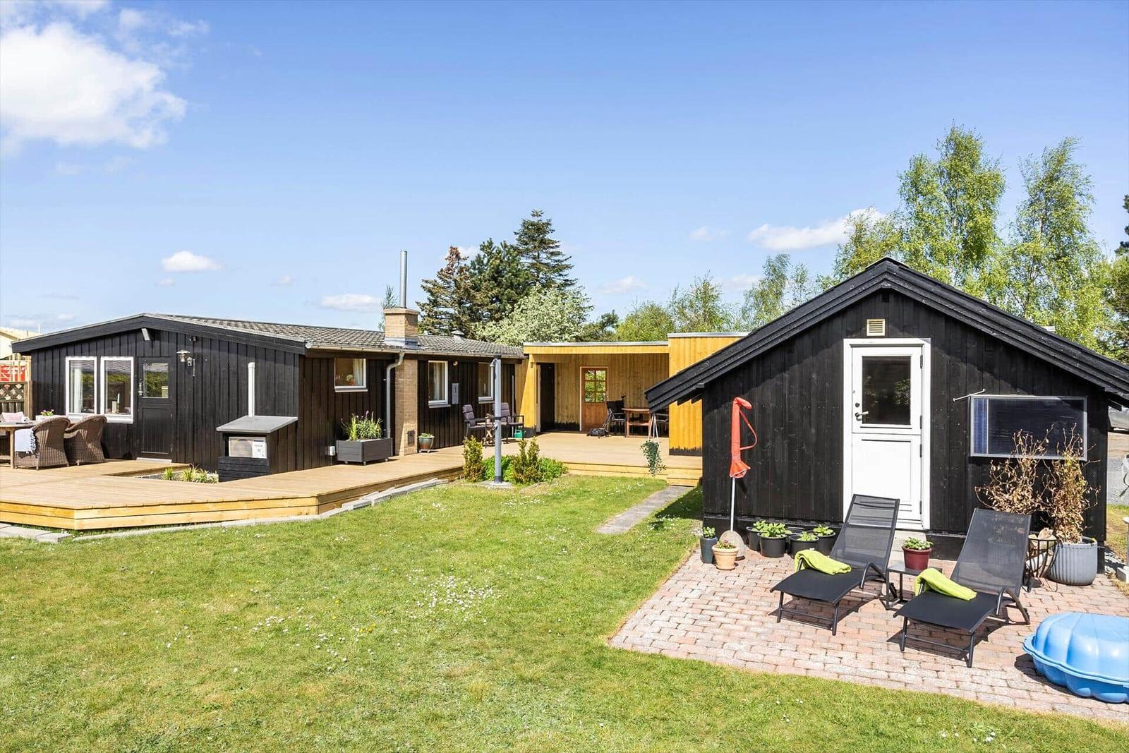 Schwarze Holzhäuser mit Terrasse und Garten. Liegestühle und Pflanzen sind vorhanden.