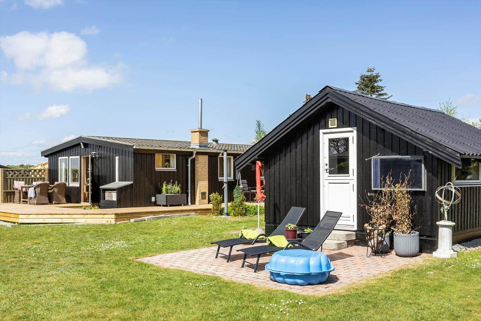 Zwei schwarze Holzhäuser mit Garten, Sonnenliegen und blauem Planschbecken.
