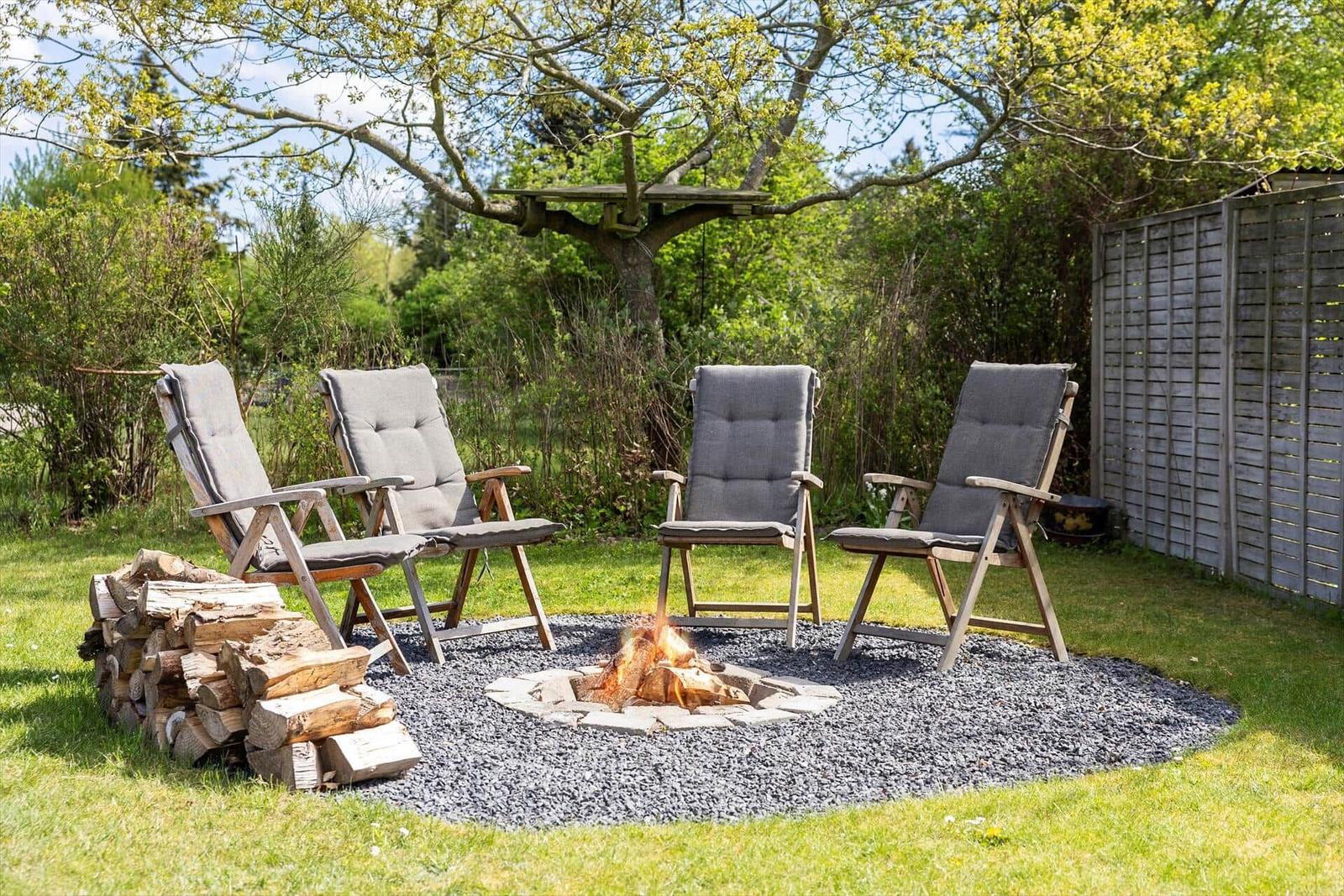 Feuerstelle mit vier Liegestühlen im Garten. Holzstapel und Baumhaus im Hintergrund.