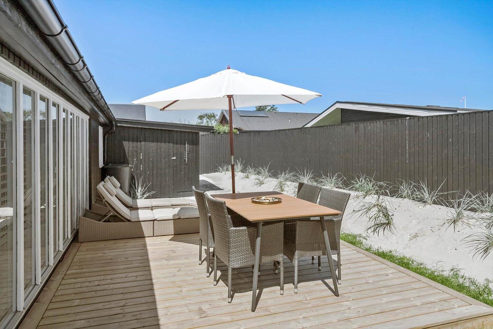 Terrasse mit Tisch, Stühlen und Sonnenschirm neben Haus mit Holzdecke und Sandhügel.