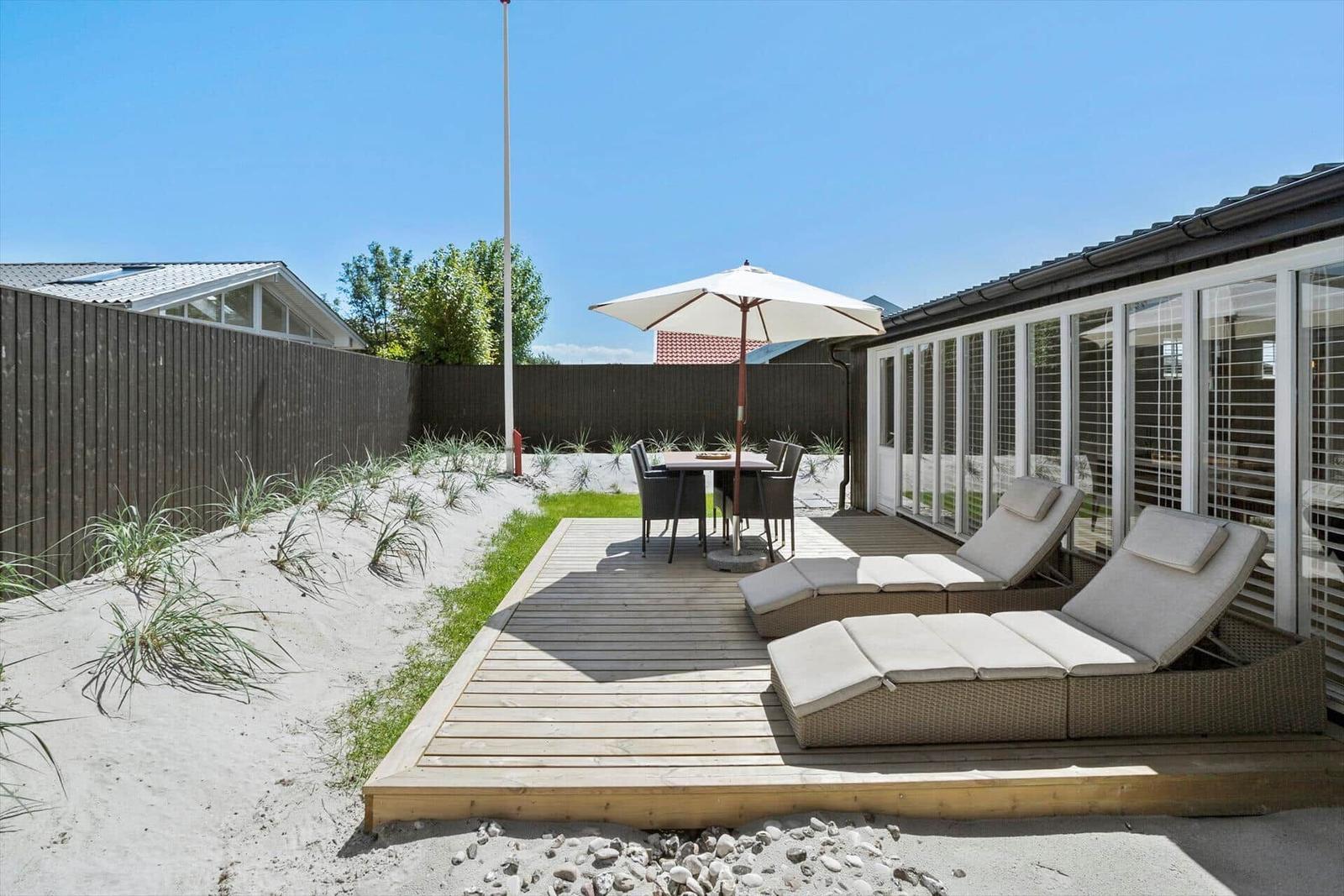 Terrasse mit Liegestühlen, Tisch und Sonnenschirm neben einem Haus mit Fenstern.