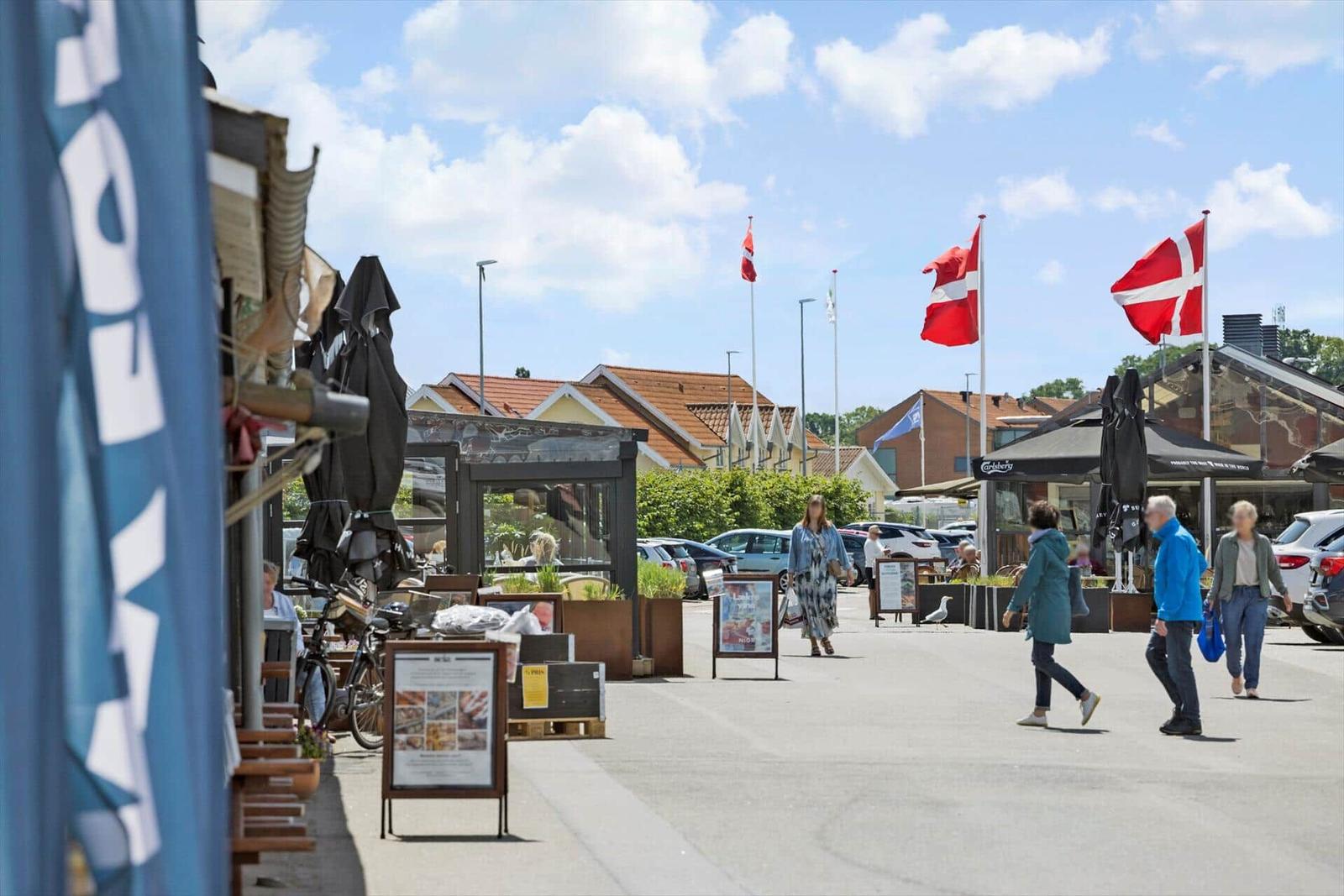 Straßenszene mit Flaggen, Geschäften und Passanten unter blauem Himmel.