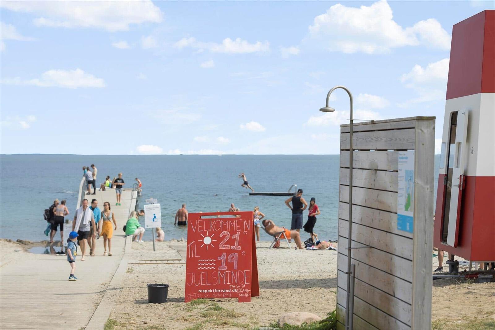 Strand mit Badesteg, Menschen und rotem Willkommensschild.