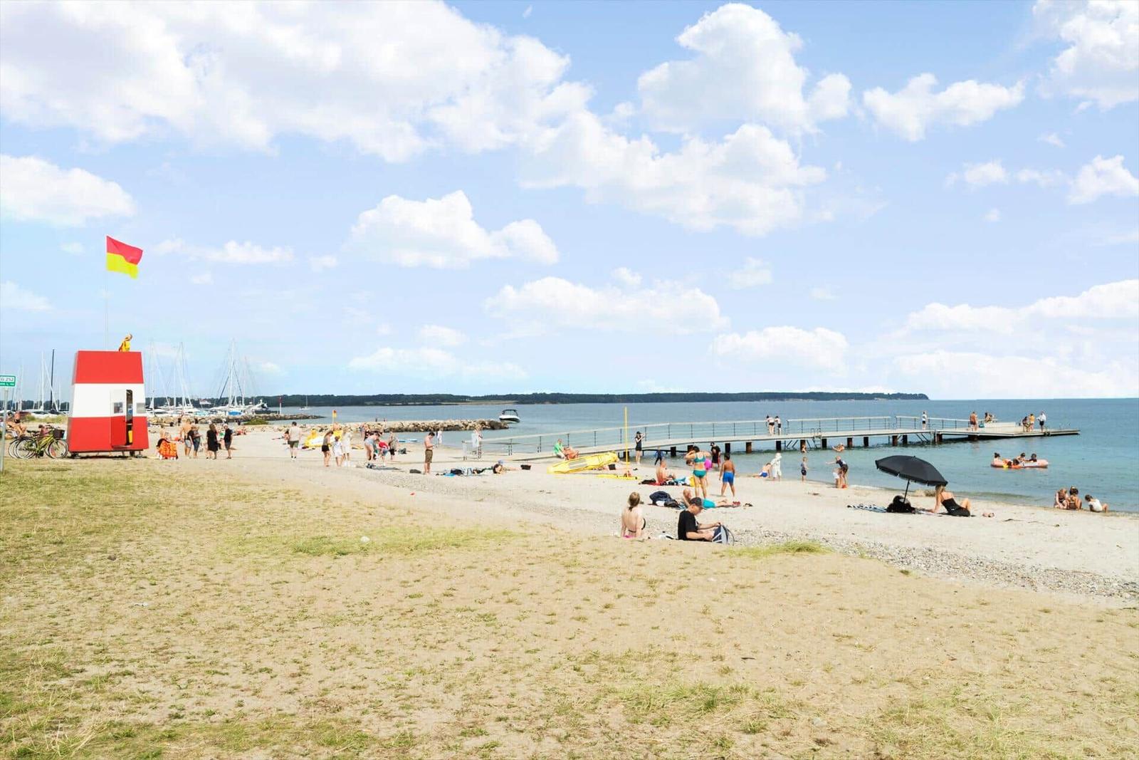 Strand mit Steg, Sonnenbadenden und Segelyachten im Hintergrund.