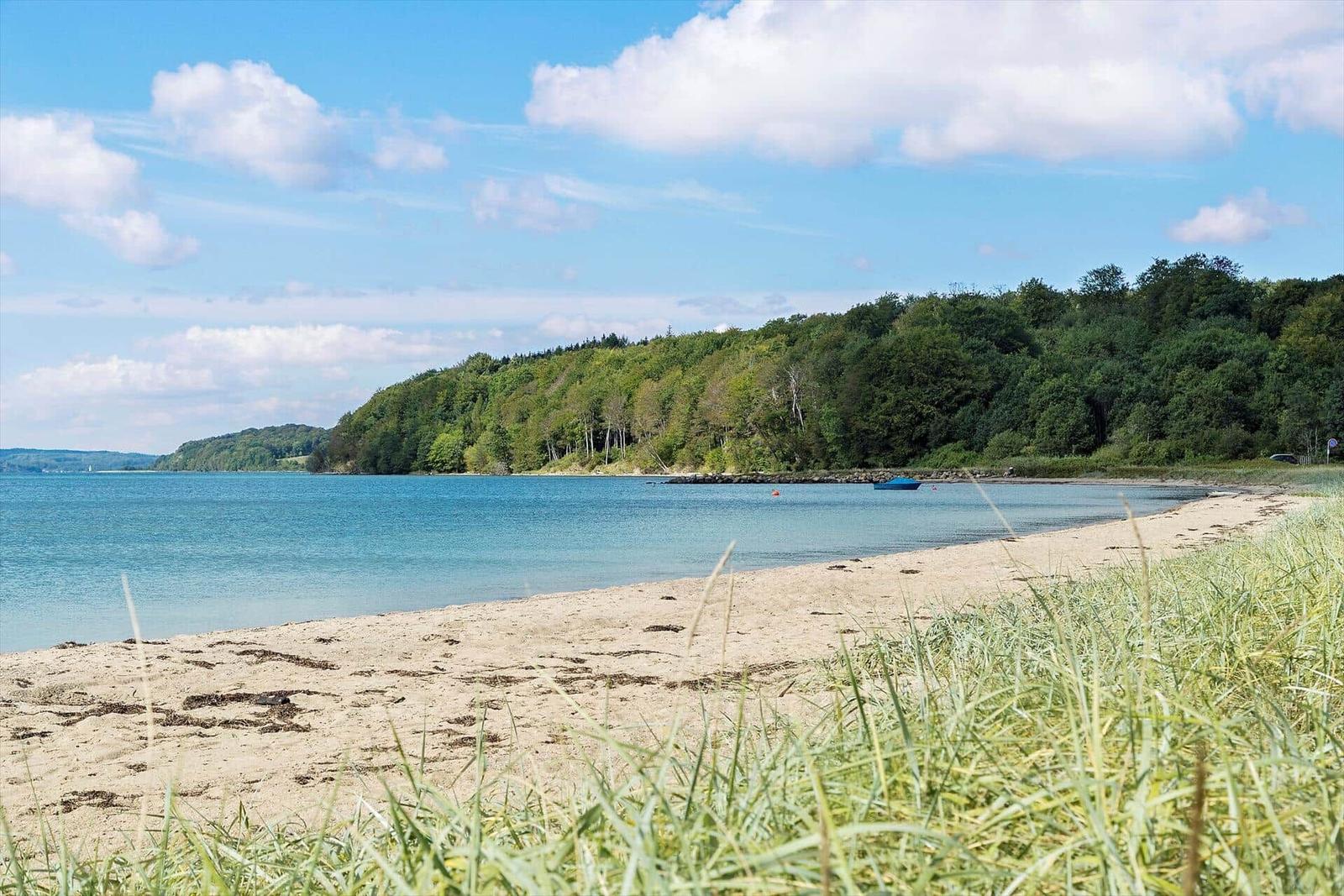 Strand mit Sand, Gras und blauem Wasser unter bewölktem Himmel.
