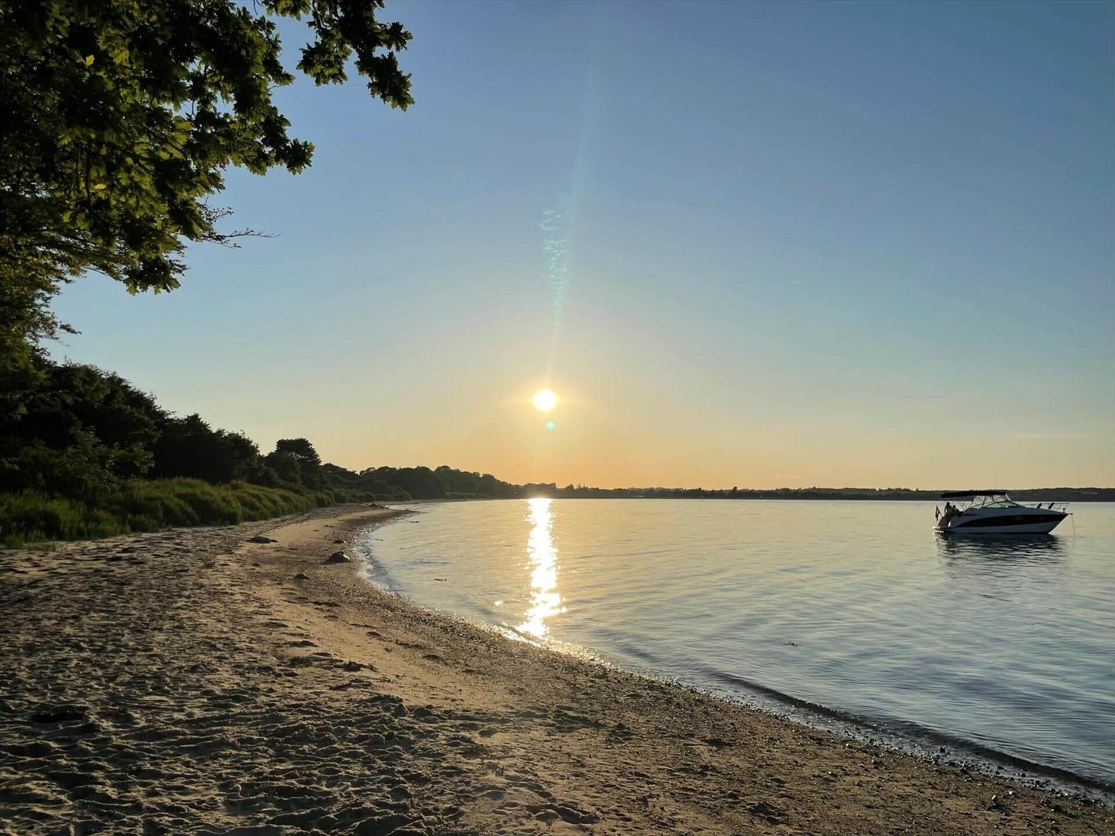 Sonnenuntergang am Strand mit Boot auf ruhigem Wasser und Bäumen im Hintergrund.