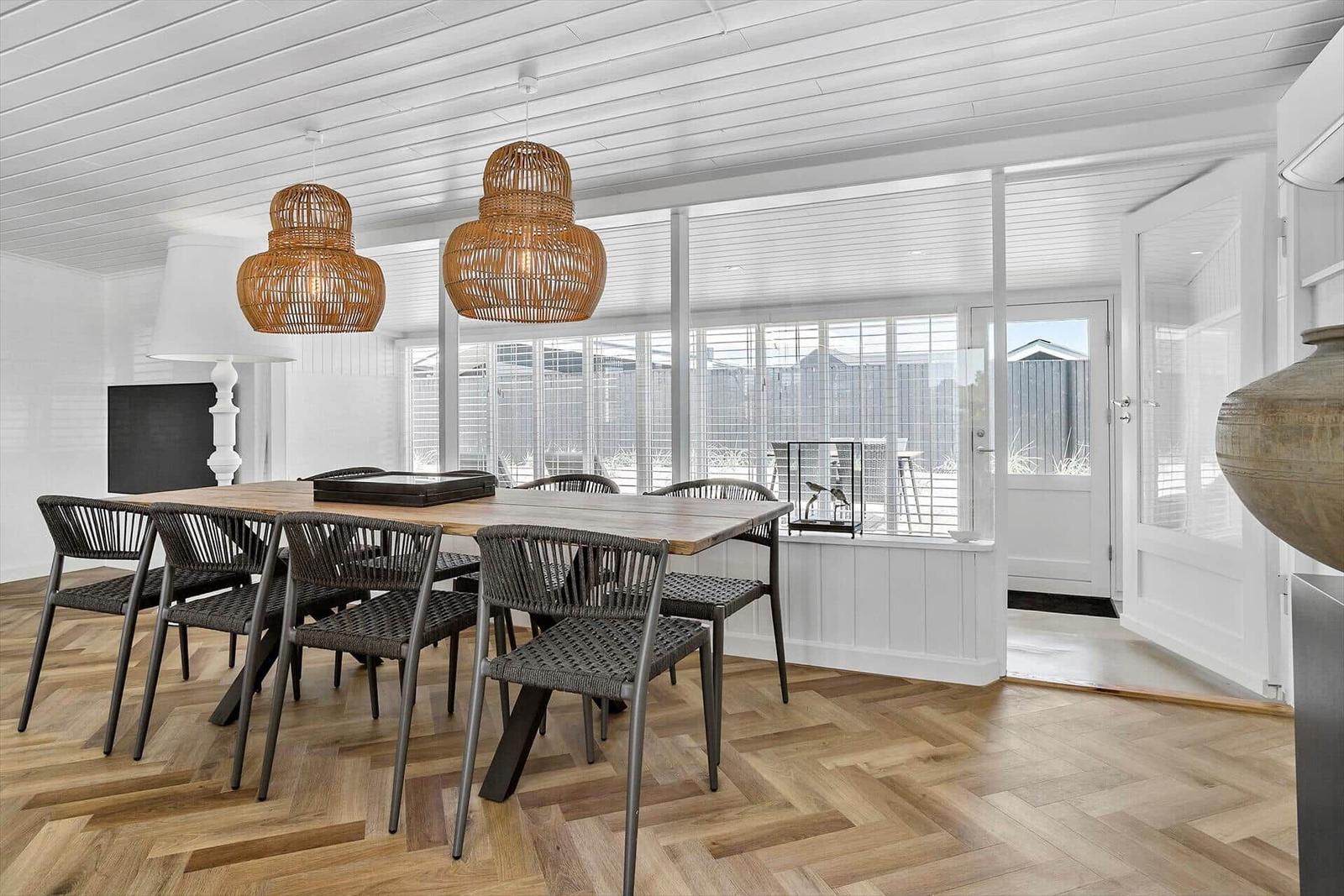 Esszimmer mit Holztisch, Stühlen und hängenden Lampen. Durchgang zur Terrasse mit Fenstern.