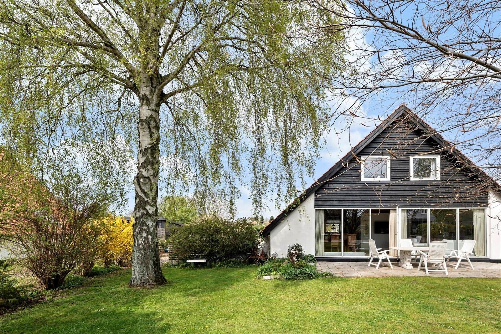 Haus mit großem Garten und Bäumen. Terrasse mit Tisch und Stühlen. Glasfront zur Aussenterrasse.